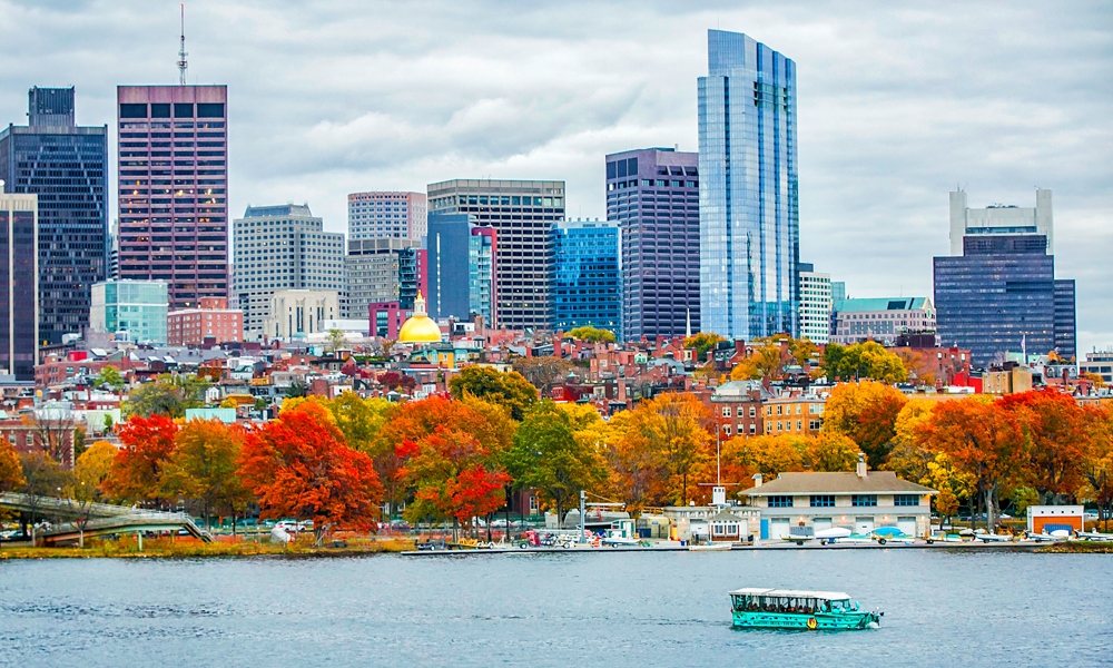 View of Boston from afar with buildings and autumn trees with Boston Duck Tour in the water.