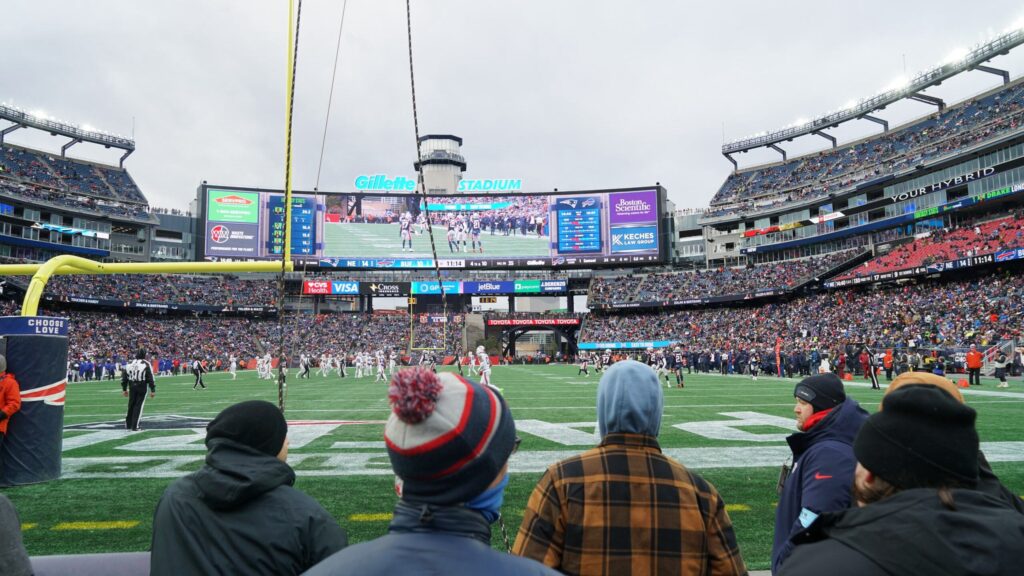 Fans with their back to the camera looking toward the stadium. One can see the scoreboard, players on the field, and crowd in the background.